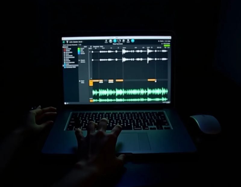 close-up of laptop screen displaying a multi-track audio session, waveform layers labeled, performer's hand resting on t