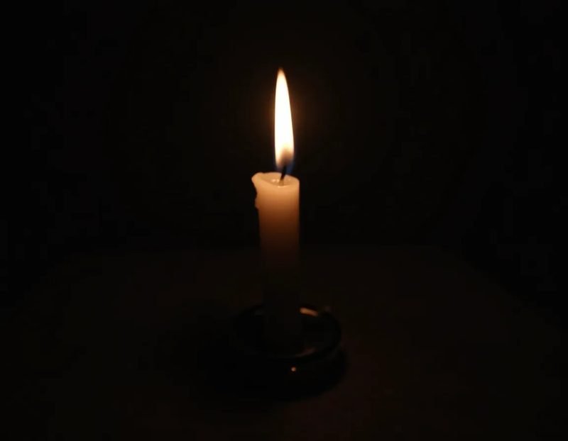 Close-up of a single taper candle on a dark tabletop, flame slightly bent