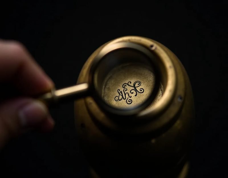 Close-up of a hand holding a magnifying loupe over engraved initials on the rim of an antique brass object